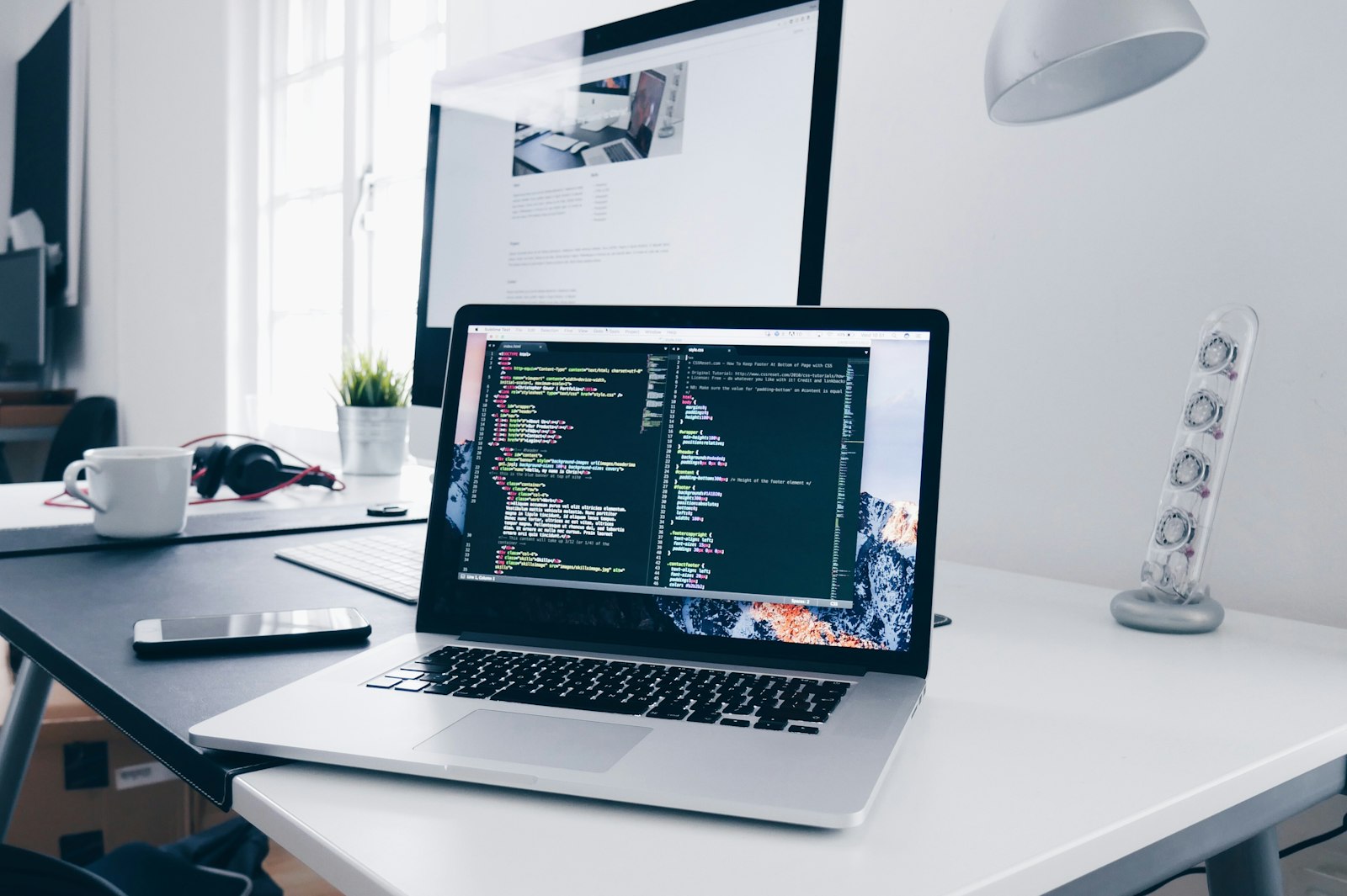 White desk with open laptop displaying code, desktop monitor, coffee mug, headphones and small plant in bright natural light